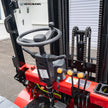 Operator station of a 6,500 lb 60V electric forklift showing steering wheel, three hydraulic control levers for mast lift and tilt, key ignition switch, red emergency stop button, and integrated instrument panel for warehouse material handling.