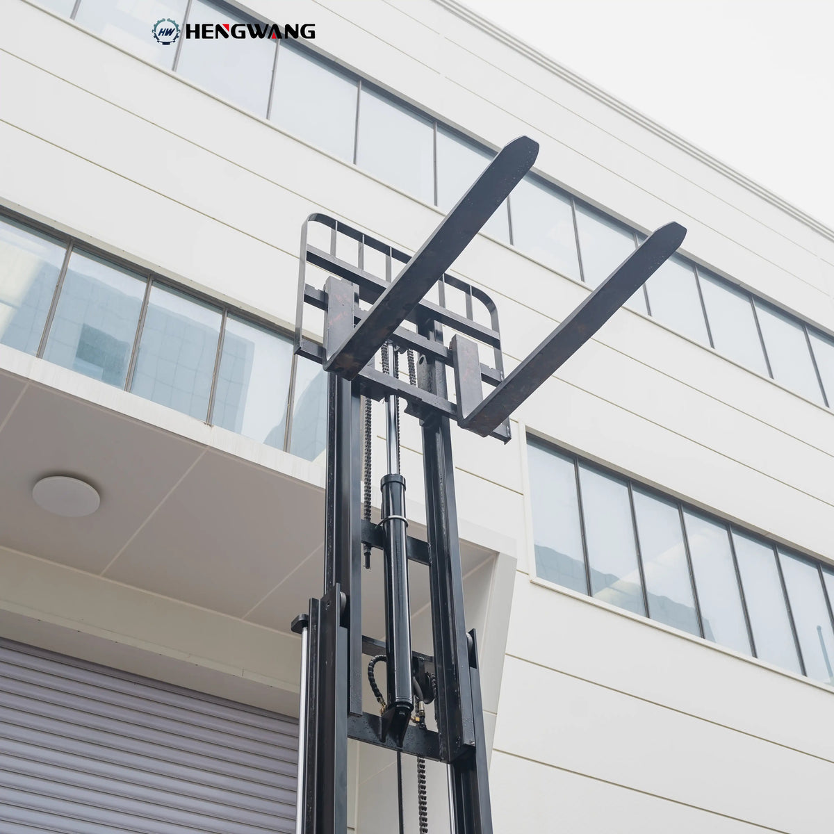 Close-up low-angle shot of 48-inch steel pallet forks on the carriage of a 6,500 lb electric forklift, showing lift chains, hydraulic cylinder, and welded mast rails for heavy warehouse load handling.