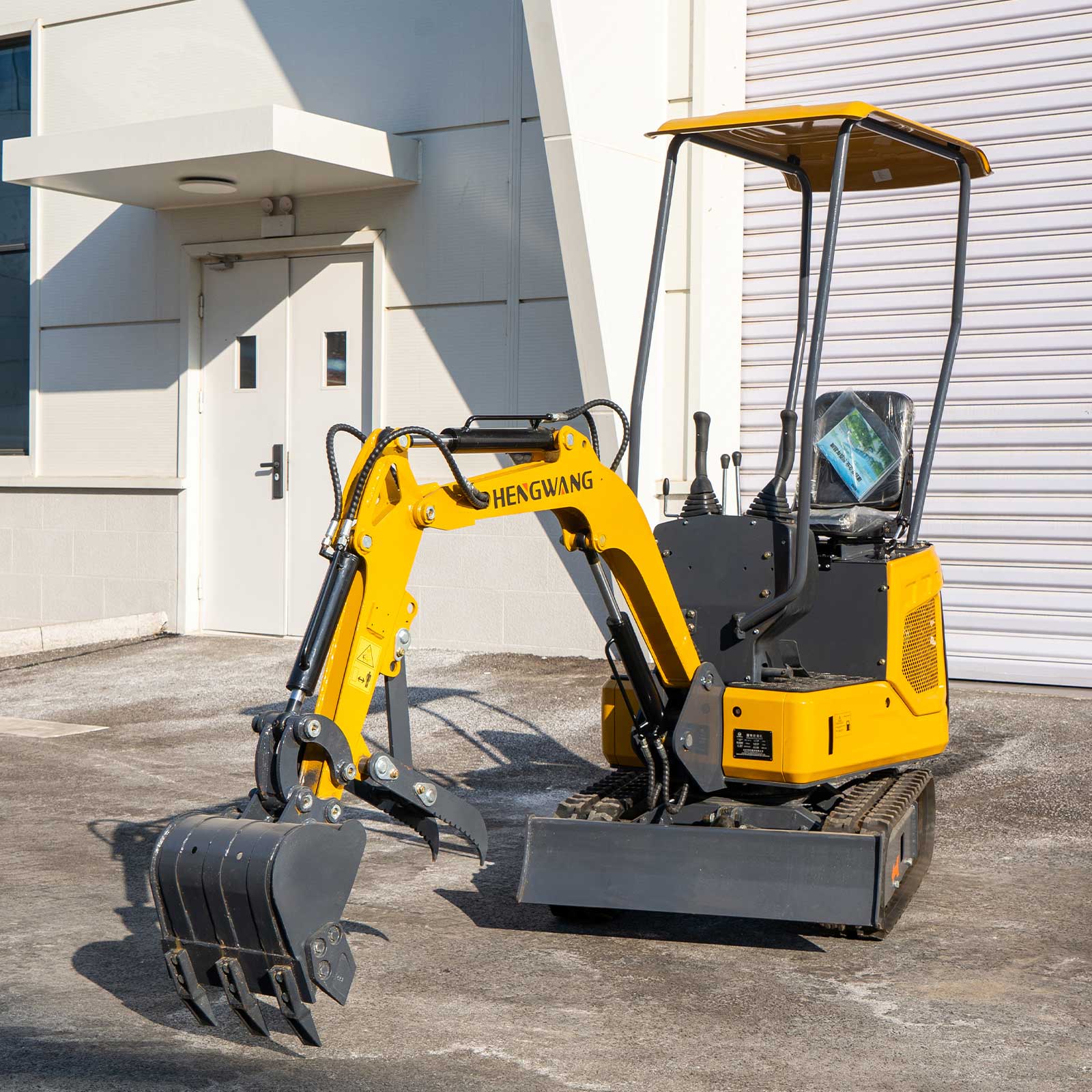 Full side front shot of HENGWANG compact mini crawler excavator fitted with digging bucket and ripper claw, rubber tracks and sun shade roof, small digger parked outside factory warehouse