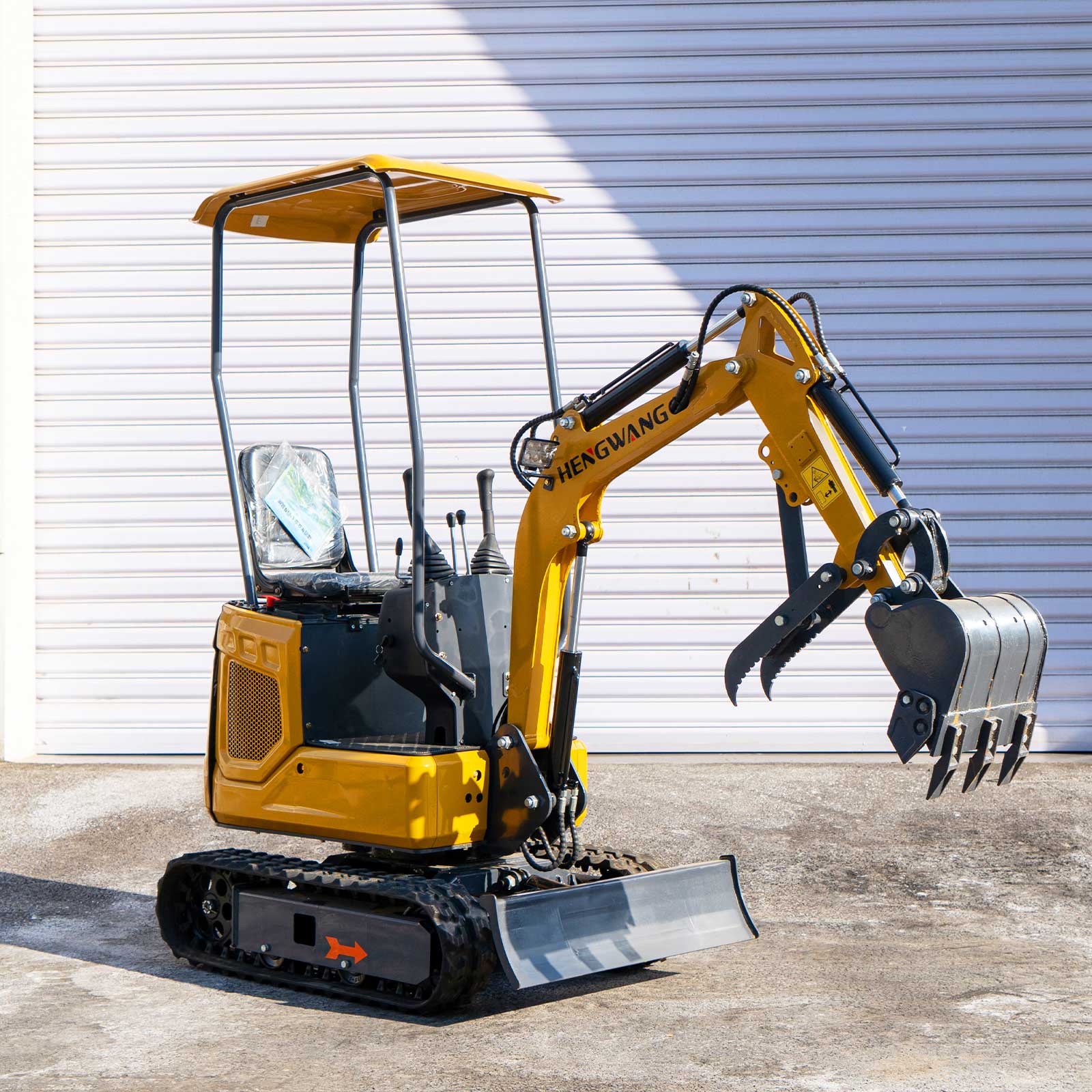 Right side full view of HENGWANG compact mini crawler excavator with digging bucket and ripper claw, rubber tracks, sun canopy roof, small digger in front of metal rolling shutter warehouse