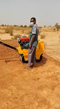 Worker standing beside yellow walk behind double drum vibratory road roller on outdoor soil construction jobsite
