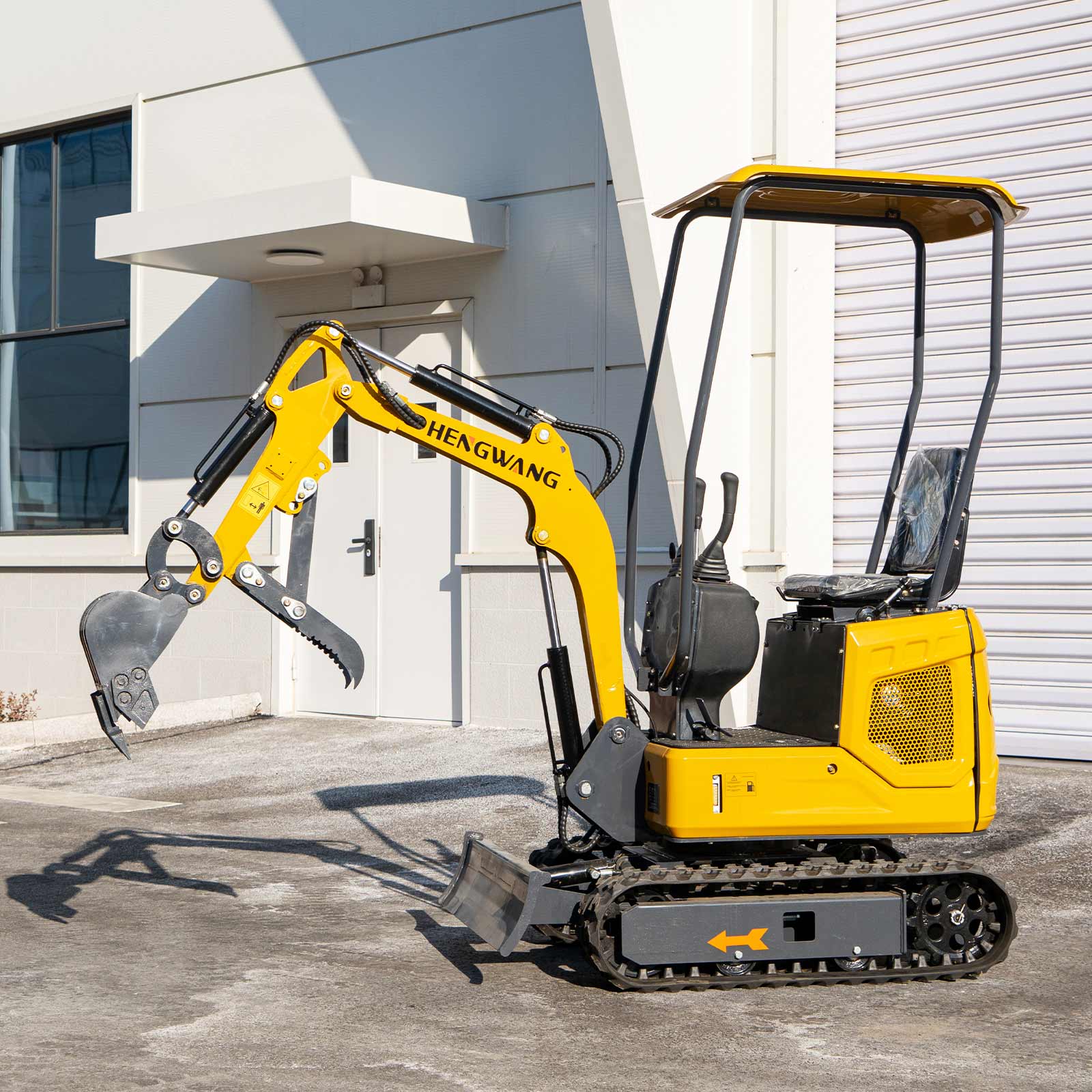 Left side profile of HENGWANG compact mini crawler excavator equipped with digging bucket and ripper attachment, lightweight rubber tracks and sun canopy, small digger parked outside modern factory building